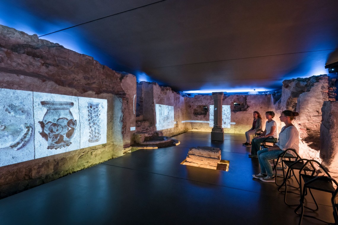 Drei Frauen auf Stühlen verfolgen Multimediashow im Schauraum Erasmuskapelle. Auf dem alten Mauern zu sehen sind Projektionen von Funden (Gefäßen).