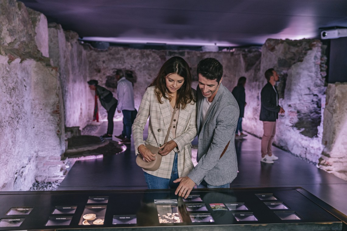 Paar mittleren Alters an Ausstellungstisch mit unterschiedlichen Funden im Schauraum Erasmuskapellen. Im Hintergrund betrachten mehrere Menschen unterschiedliche Stellen des alten Gemäuers.